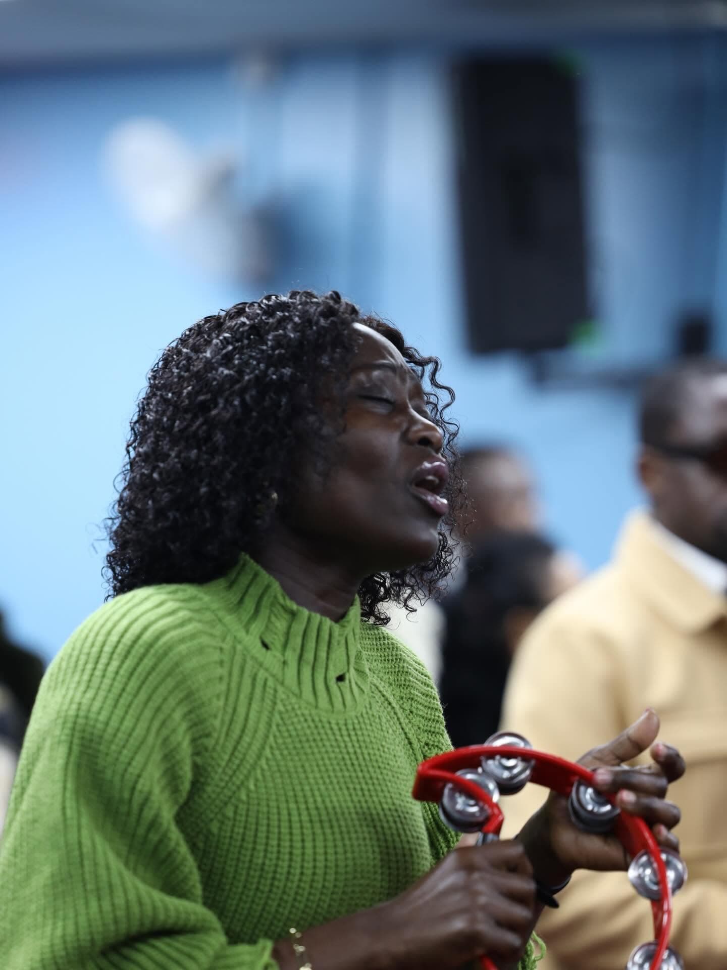 A woman in a green jumper singing and playing a red tambourine during praise